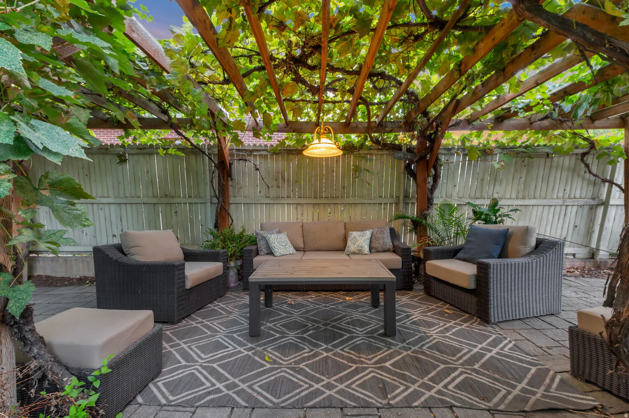 Paved patio area featuring a wooden pergola with mature vines, a slatted privacy fence, and an overhead light fixture