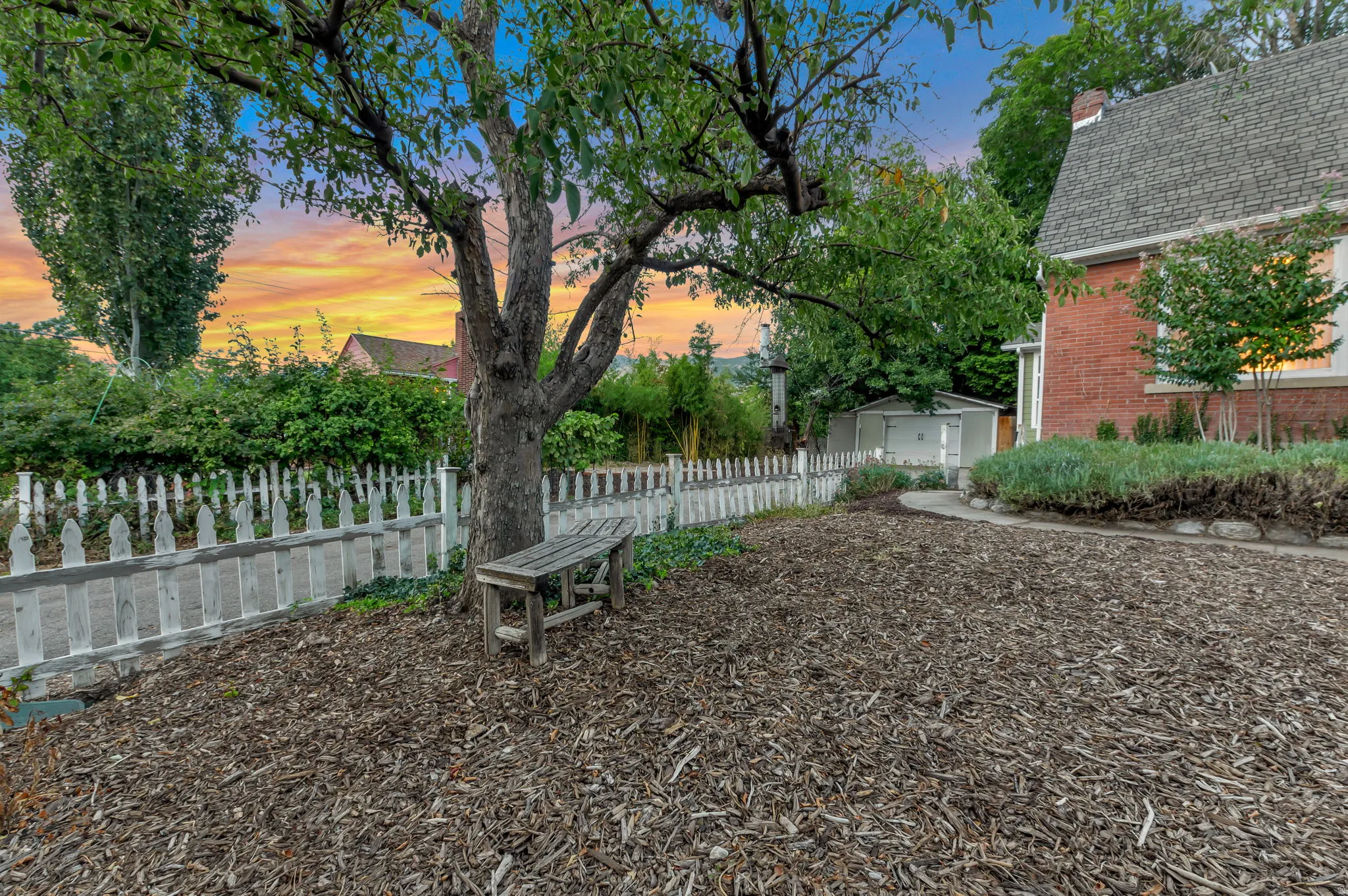 Brick exterior residence featuring a gabled roof, white picket fence, mature tree, and a storage shed