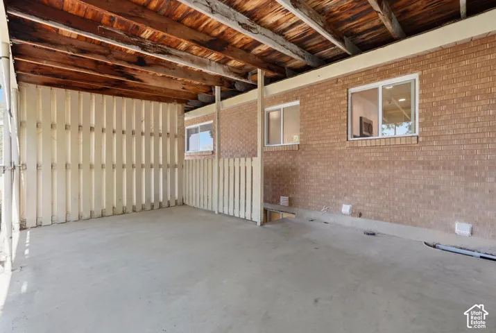 Covered concrete patio featuring an exposed wood beam ceiling, brick exterior wall, and white trim windows