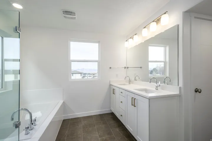 Dual vanity with quartz-style countertops and brushed brass hardware, large mirrored wall, separate soaking tub, walk-in glass shower, and rectangular window