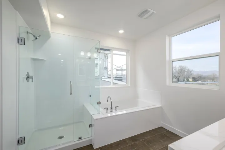 Glass-enclosed shower with a built-in shelf, deep soaking tub with deck-mounted faucet, and multiple windows providing natural illumination