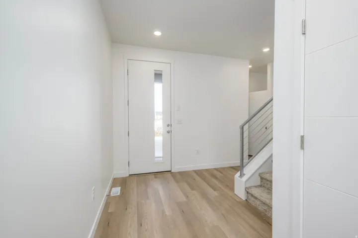 Recessed ceiling lighting, wood-finish flooring, white walls, modern entry door with vertical glass insert, and a staircase with cable railing
