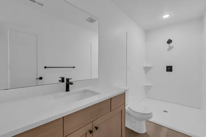 Wood-finish flooring, single vanity with light-toned countertop, built-in rectangular sink, matte black faucet, and walk-in shower with tile surround