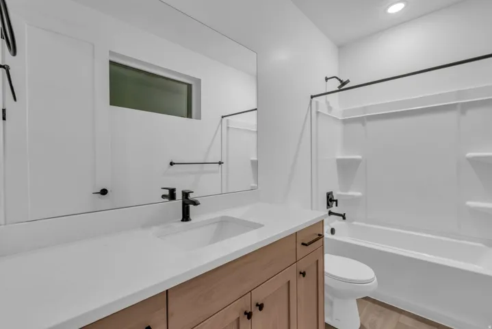 White solid surface countertop with integrated sink, wood-finish vanity with black hardware, matte black faucet, and white shower/tub combination with black fixtures
