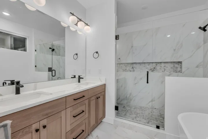 Dual vanity with rectangular undermount sinks and matte black fixtures, wood-finish cabinetry, frameless glass shower with marble-look tile surround and mosaic detail, and a built-in shower niche