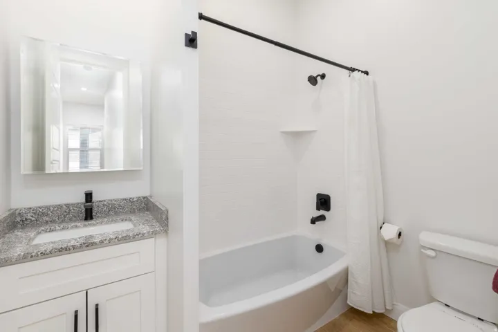 White subway tile shower surround with integrated shelf, white bathtub with matte black fixtures, white vanity with granite countertop, integrated sink, and matte black faucet, and a wall-mounted medicine cabinet