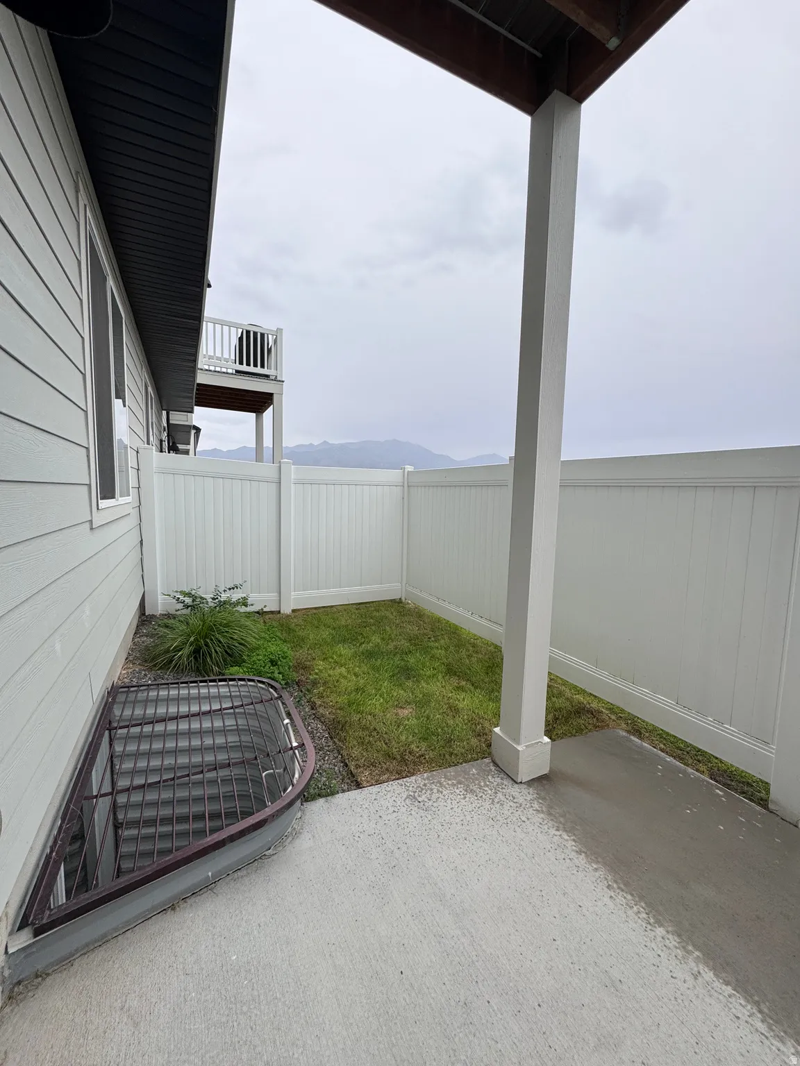 Covered patio with a concrete surface, enclosed by a white privacy fence, featuring a small lawn area and an egress window well