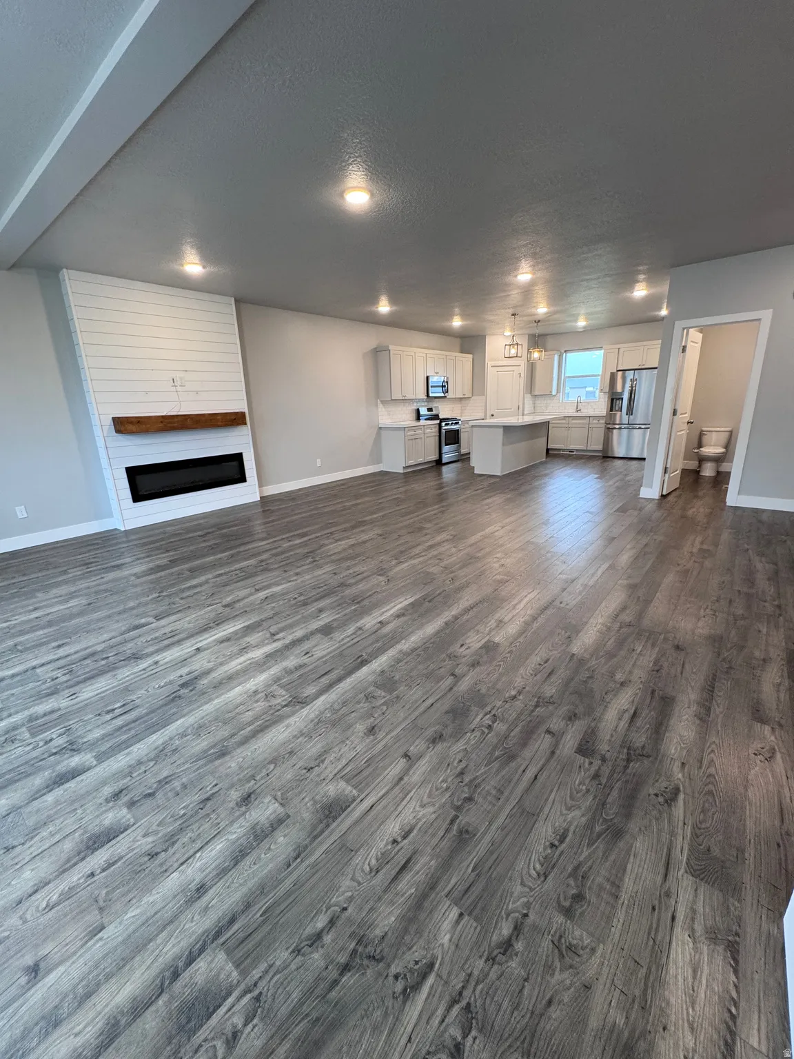 Open-concept living area with plank wood-finish flooring, a shiplap fireplace with a wood mantle, recessed lighting, and a kitchen featuring a center island and stainless steel appliances