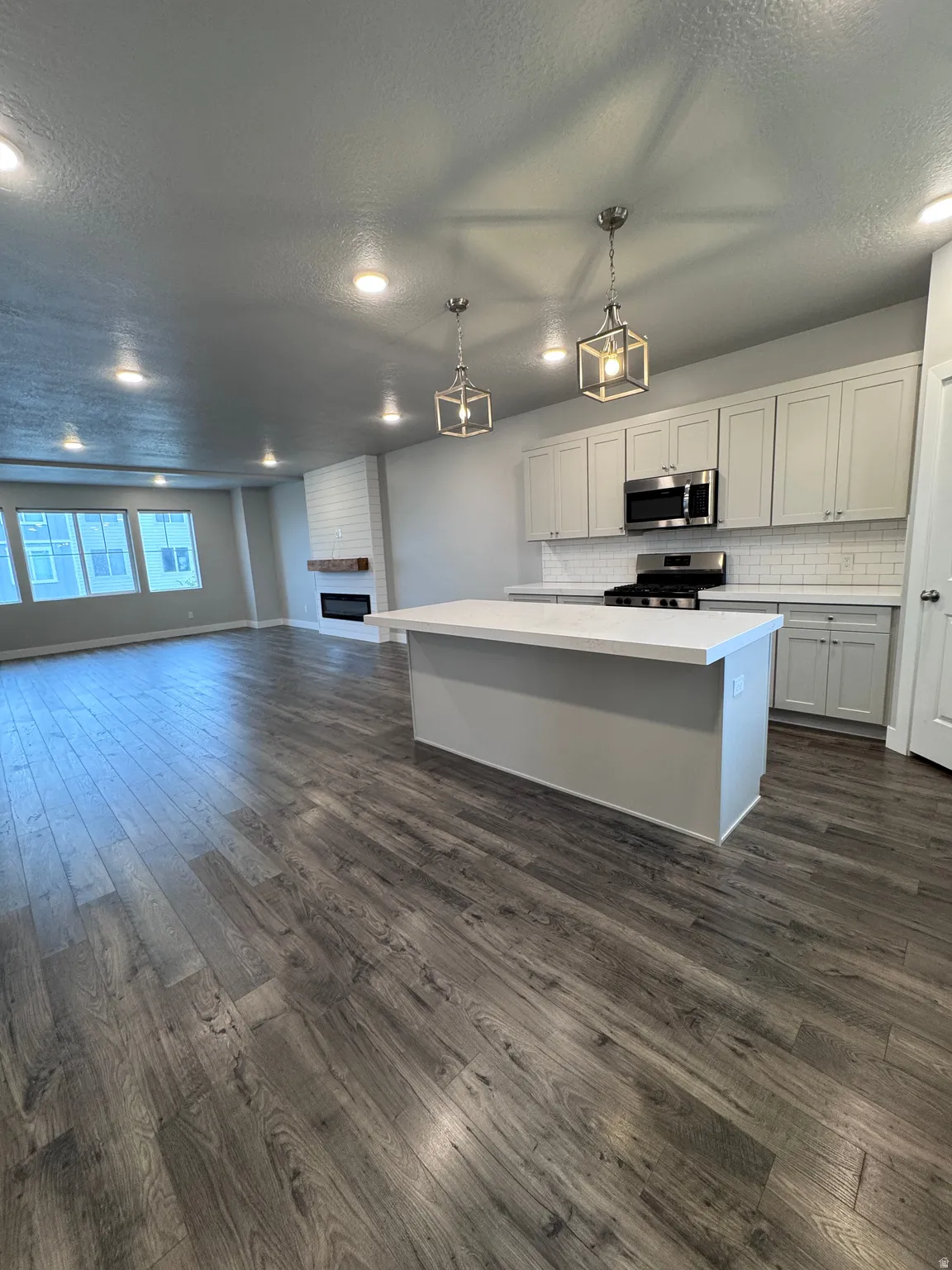 Open-concept living space with wood-finish flooring, recessed lighting, a kitchen island with a solid surface countertop, white subway tile backsplash, and a fireplace with a wood mantle