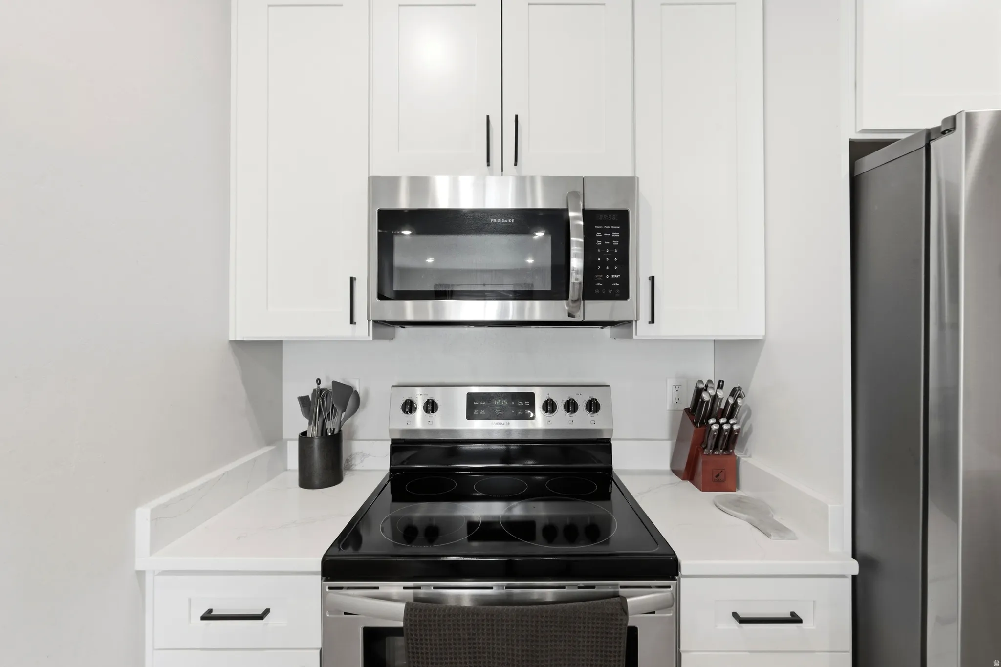White cabinetry with matte black hardware, stainless steel microwave, stainless steel range, and light-toned countertops