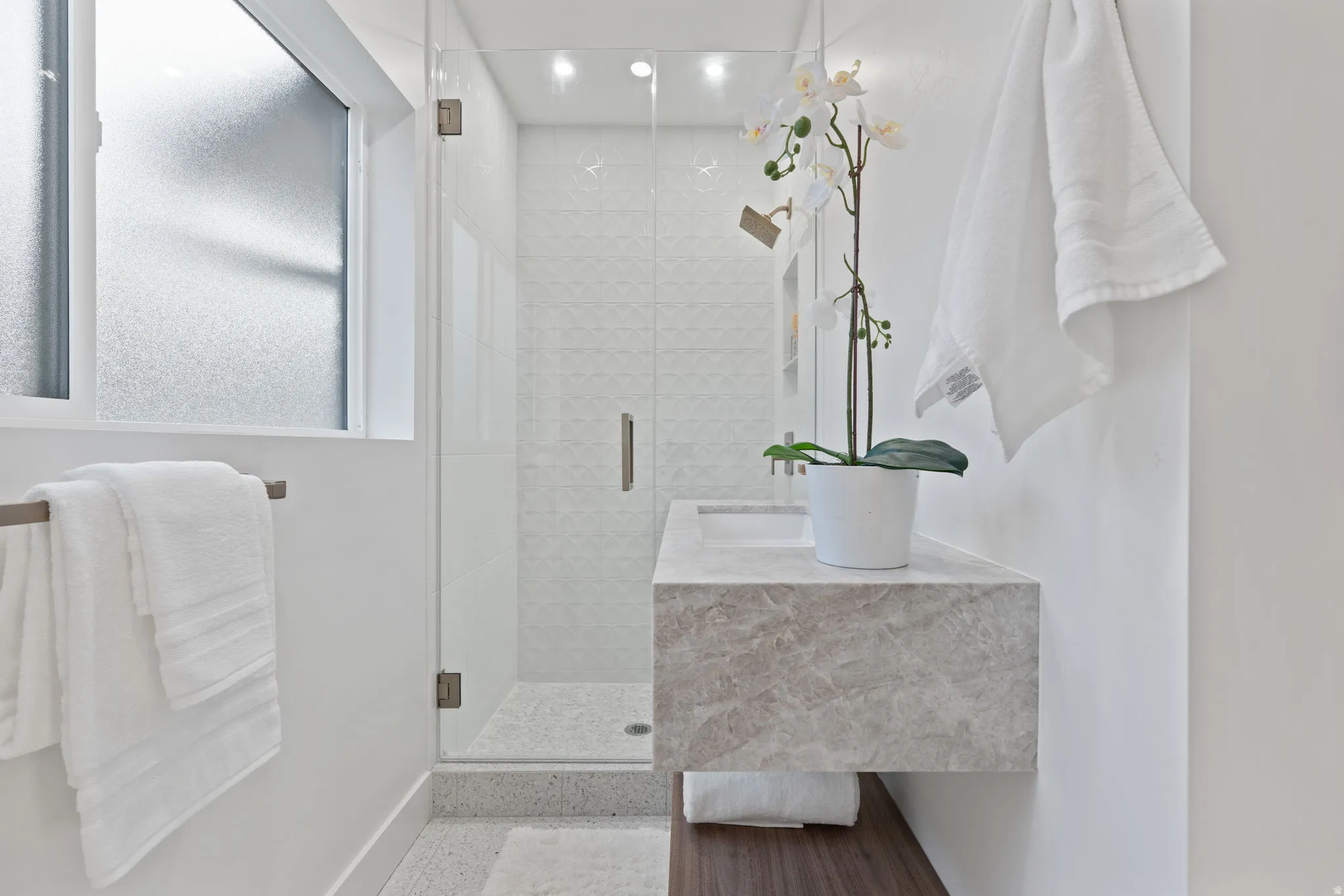 Floating vanity with integrated basin, frameless glass shower enclosure, textured shower wall tile, and frosted window