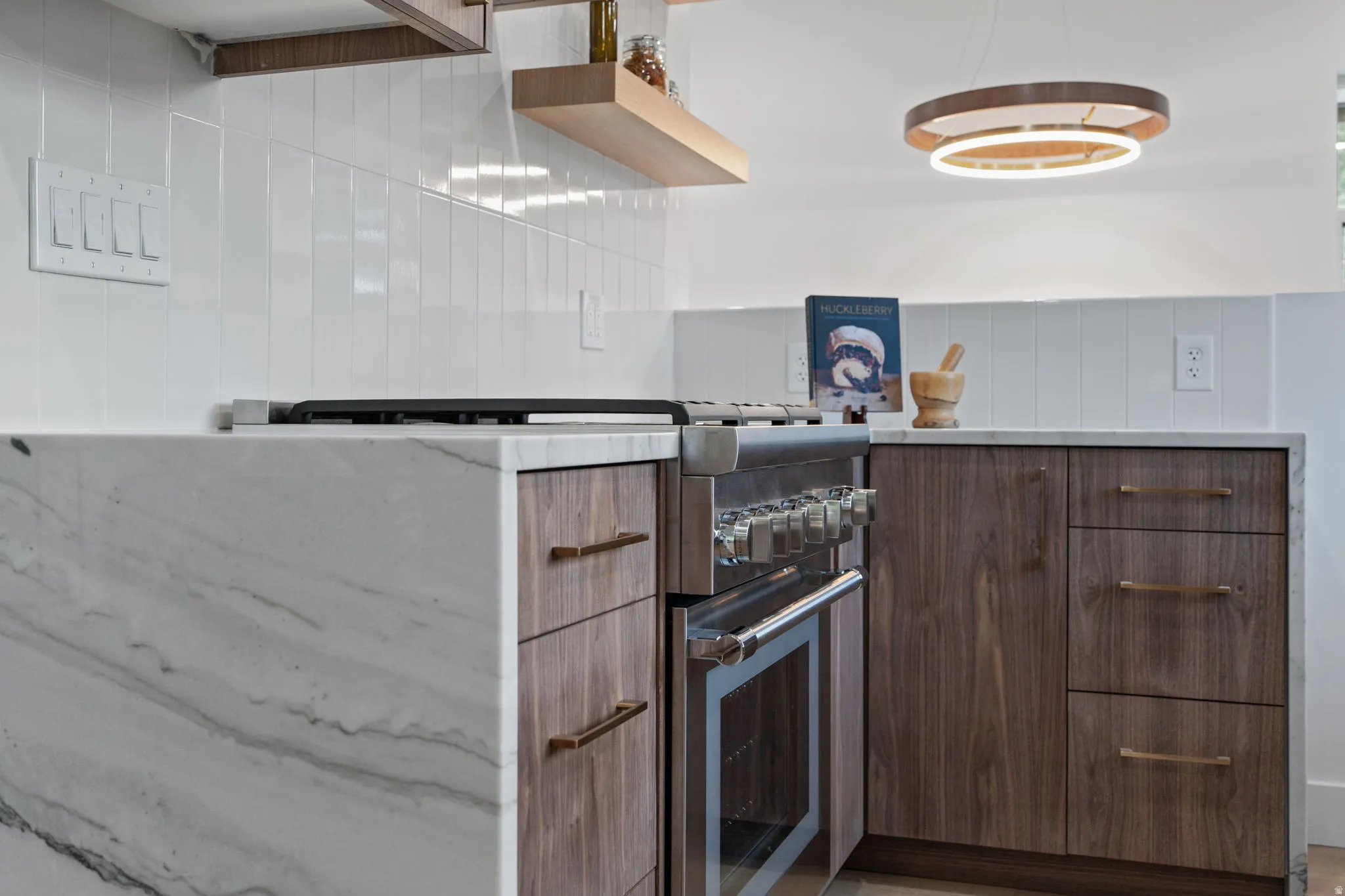 Stainless steel range, wood-finish cabinetry, waterfall-edge countertop, white tile backsplash, and a contemporary circular pendant light