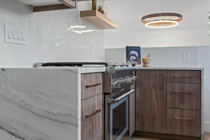 Stainless steel range, wood-finish cabinetry, waterfall-edge countertop, white tile backsplash, and a contemporary circular pendant light