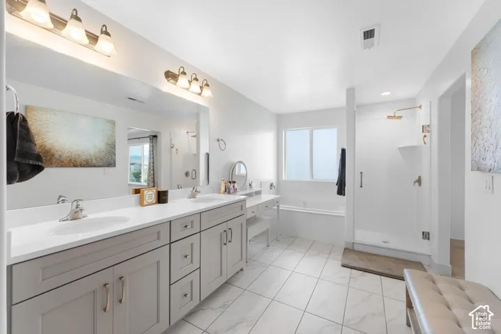Dual sink vanity with light gray cabinetry and brushed nickel hardware, a separate built-in soaking tub, a glass-enclosed shower with a rain shower head, and large format floor tiling