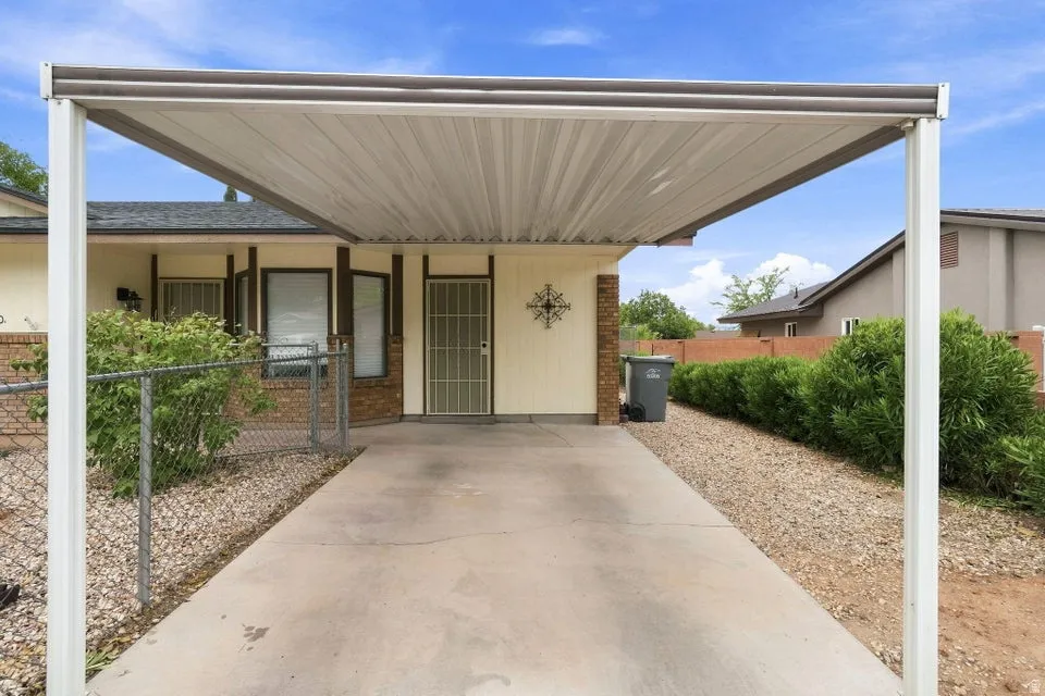 Carport structure providing covered parking, concrete driveway, decorative security door, brick facade accents, and mature landscaping