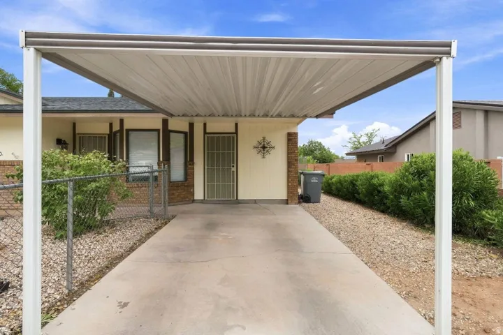Carport structure providing covered parking, concrete driveway, decorative security door, brick facade accents, and mature landscaping