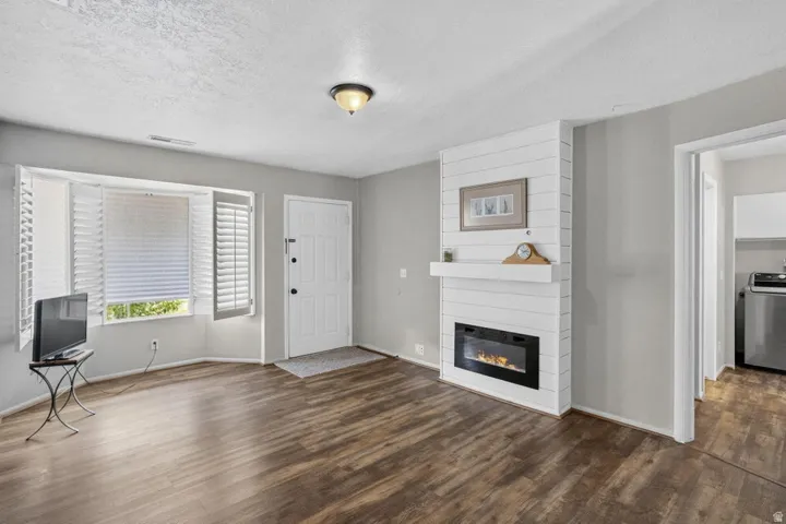 Wood-finish flooring, bay window with plantation shutters, front entry door, and a shiplap-accented fireplace with a mantel