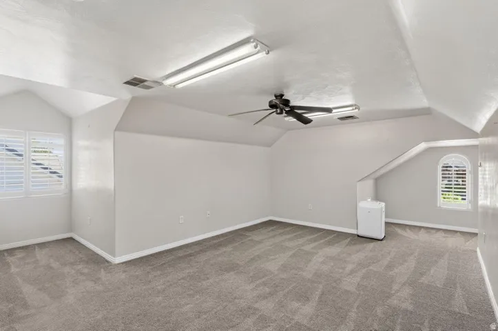 Sloped ceilings, patterned carpet flooring, ceiling fan, and window with plantation shutters