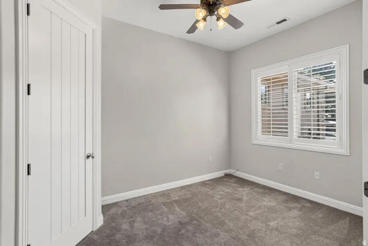 Recessed window with plantation shutters, ceiling fan with integrated lighting, paneled interior door, and grey carpet flooring