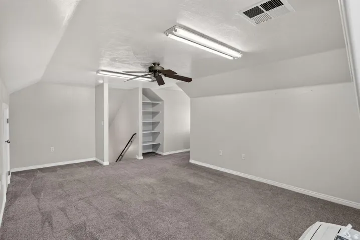 Spacious room with gray carpet flooring, sloped ceiling, built-in shelving, and a ceiling fan with integrated lighting