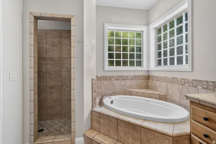 Built-in soaking tub surrounded by tile, walk-in shower with tile surround, two windows with decorative glass, and a wood-finish vanity with a stone countertop
