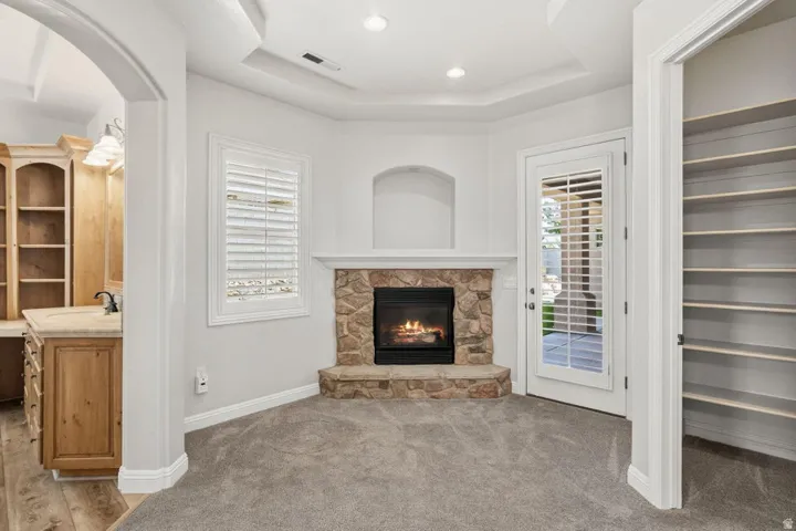 Stone-clad fireplace with hearth, tray ceiling, window with plantation shutters, glass-paneled door, and built-in shelving
