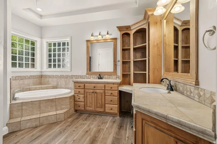 Dual wood-finish vanities with undermount sinks and tile countertops, built-in shelving, a tiled soaking tub, and wood-finish flooring