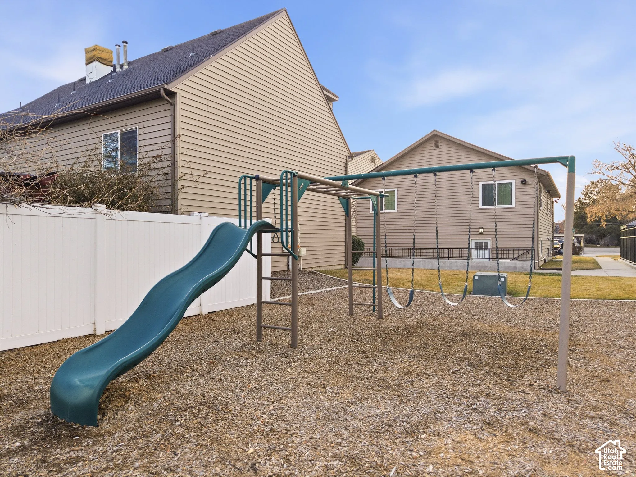 Fenced backyard with playground equipment including a slide and monkey bars, situated on a gravel surface