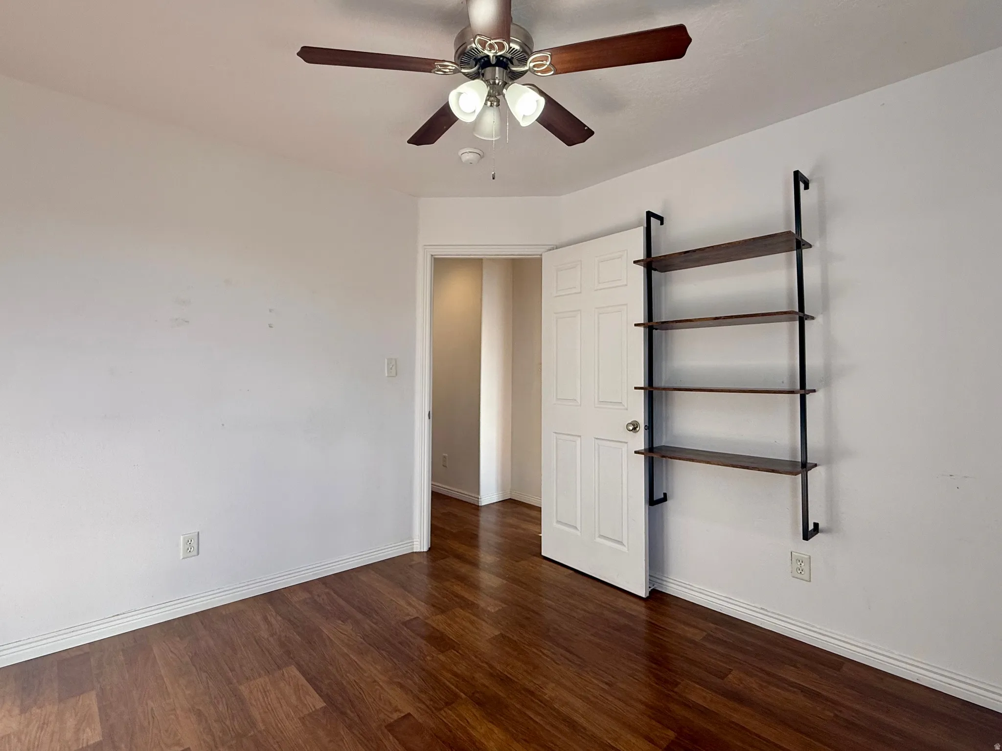 Wood-finish flooring, ceiling fan with integrated lighting, and a built-in wall shelf with dark-toned wood shelves and black metal supports