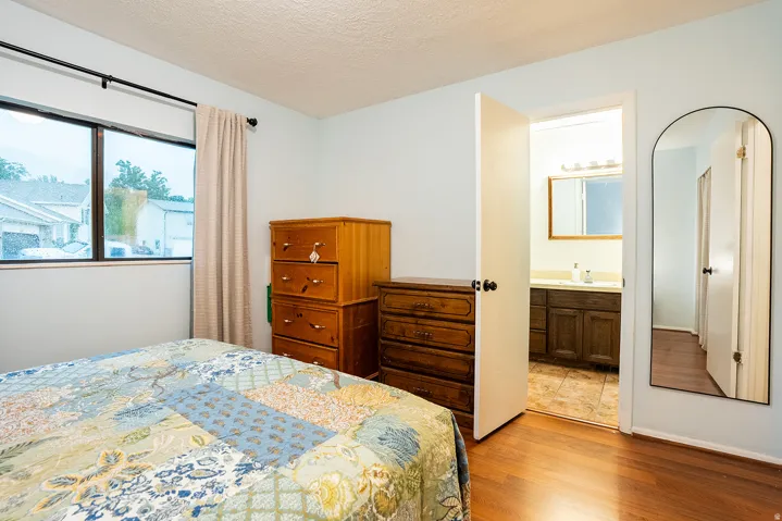 Bright primary bedroom featuring hardwood flooring, large windows with natural light, wooden dresser and nightstand storage, full ensuite bathroom with vanity and mirror, and direct access to adjoining bathroom spaces
