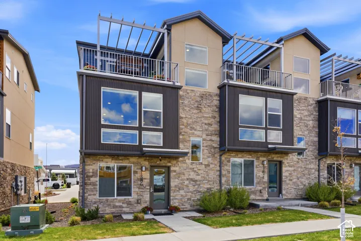 Modern three-story exterior featuring stone veneer siding, corrugated metal accents, expansive windows, and a rooftop balcony with a pergola