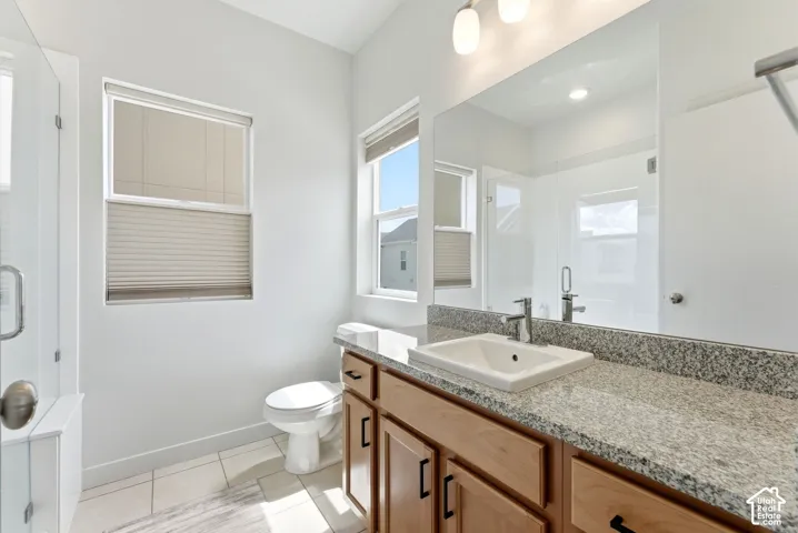 Bathroom featuring a stone-composite countertop, undermount sink with a chrome faucet, light wood cabinetry with matte black hardware, tile flooring, and a glass-enclosed shower