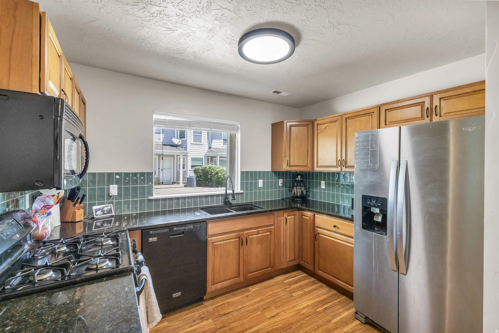 Kitchen featuring wood-finish flooring, natural wood cabinetry, dark countertops, a tile backsplash, and stainless steel appliances