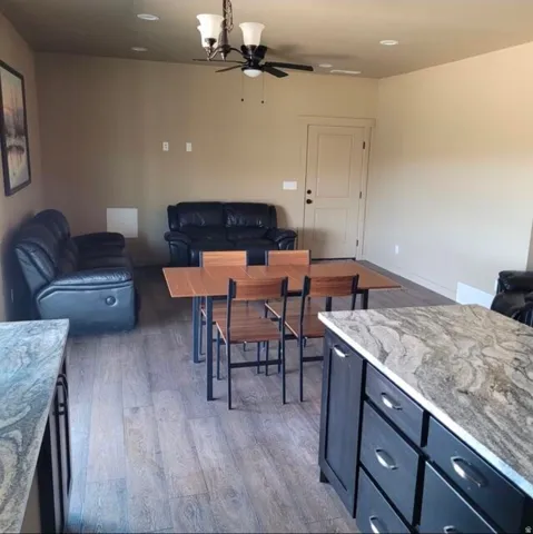 Open-concept living area featuring wood-finish flooring throughout, a dining table with seating for four, dark leather reclining seating, and a chef's kitchen with stone-finish countertops and dark cabinetry