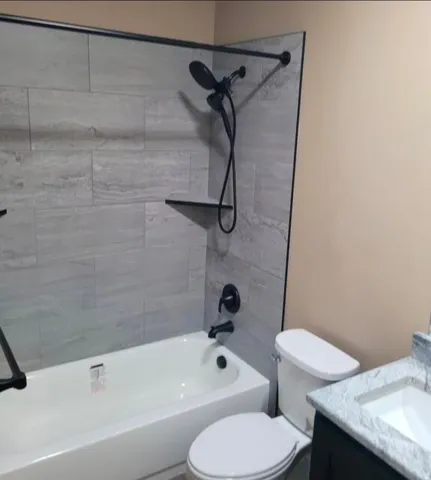 White bathtub with a matte black shower system, light-toned wall tiling, built-in shower shelf, and a white toilet