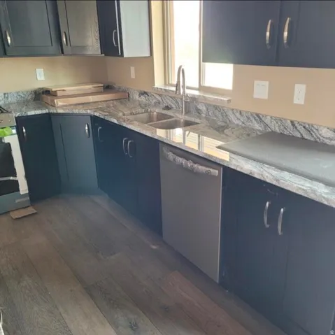 Dark wood-finish cabinetry with silver hardware, light-toned countertops with gray veining, stainless steel undermount double sink with a high-arc faucet, stainless steel dishwasher, and wood-finish flooring