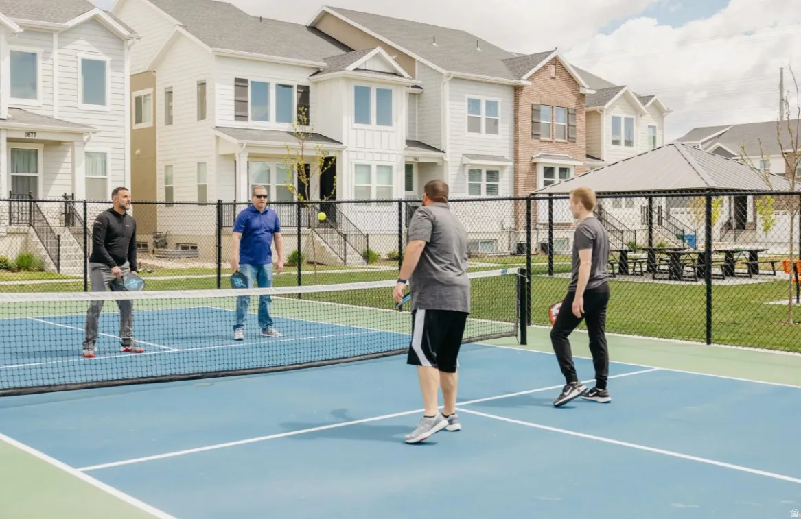 Community pickleball court featuring a blue and green playing surface, black chain-link fencing, and a nearby covered pavilion with picnic tables
