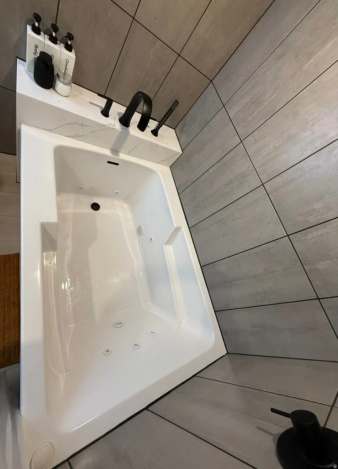 White jetted bathtub with a white marble-patterned surround, matte black fixtures, and large format grey wall and floor tiles