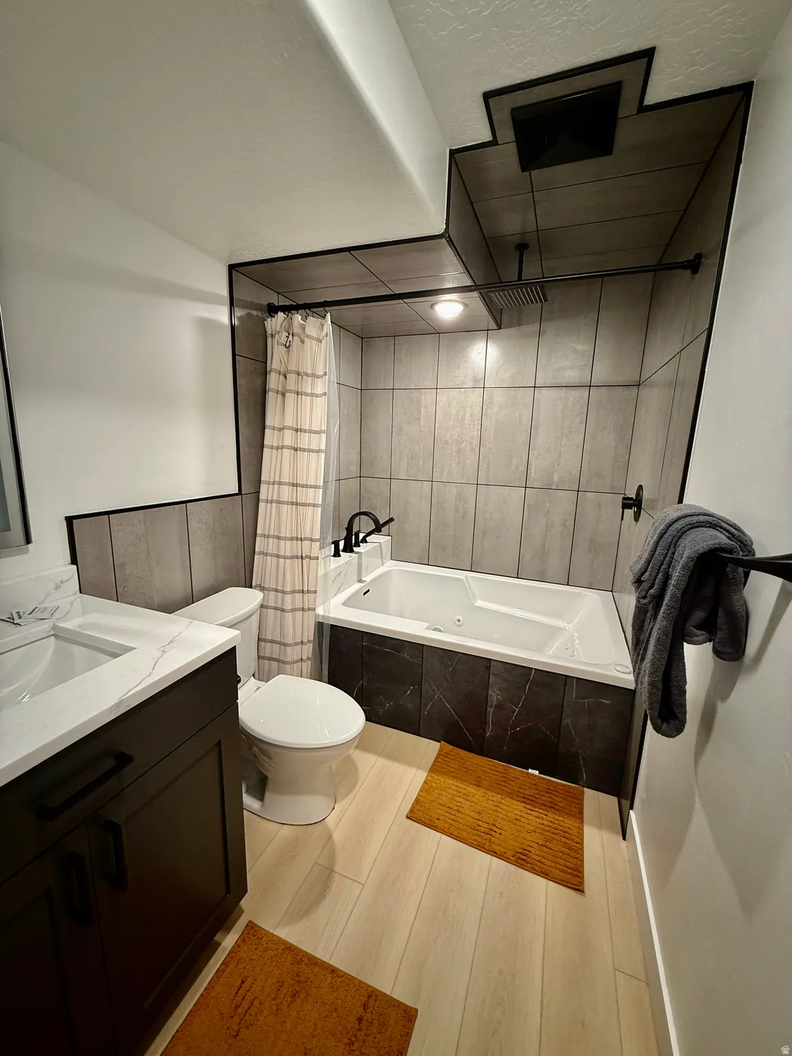 Contemporary bathroom featuring a built-in tub with textured tile surround, wood-finish flooring, a vanity with a white countertop, and modern black fixtures