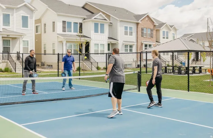 Community pickleball court featuring a blue and green playing surface, black chain-link fencing, and a nearby covered pavilion with picnic tables
