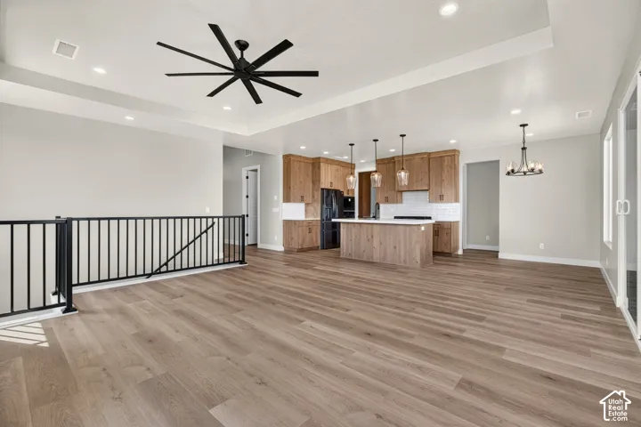 Open-concept living area with wood-finish flooring, a kitchen featuring custom wood cabinetry and white countertops, a large kitchen island with pendant lighting, a black metal staircase railing, and a tray ceiling with recessed lighting
