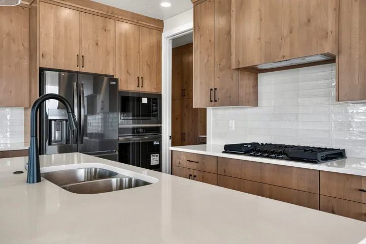 Wood-finish cabinetry with matte black hardware, stainless steel appliances, white subway tile backsplash, light-toned countertops, and an undermount double basin sink with a matte black faucet