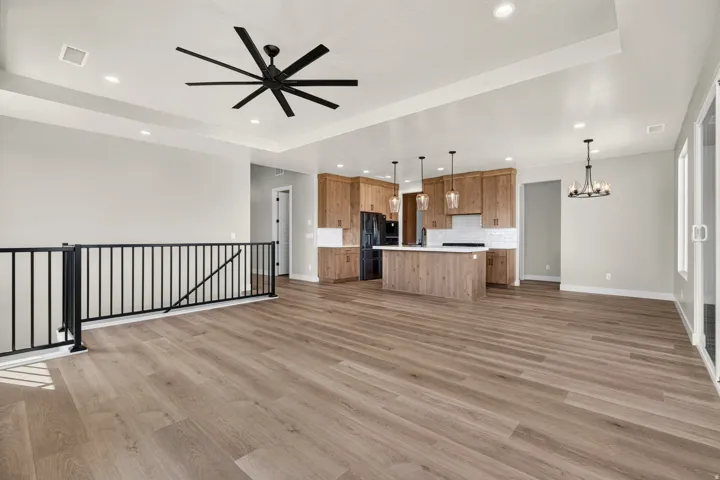 Open-concept living area with wood-finish flooring, a kitchen featuring custom wood cabinetry and white countertops, a large kitchen island with pendant lighting, a black metal staircase railing, and a tray ceiling with recessed lighting