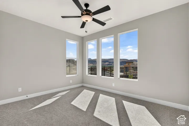 Three large picture windows, grey carpeting, neutral wall paint, and a ceiling fan with integrated lighting