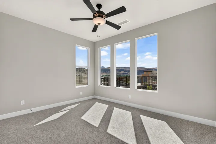 Three large picture windows, grey carpeting, neutral wall paint, and a ceiling fan with integrated lighting