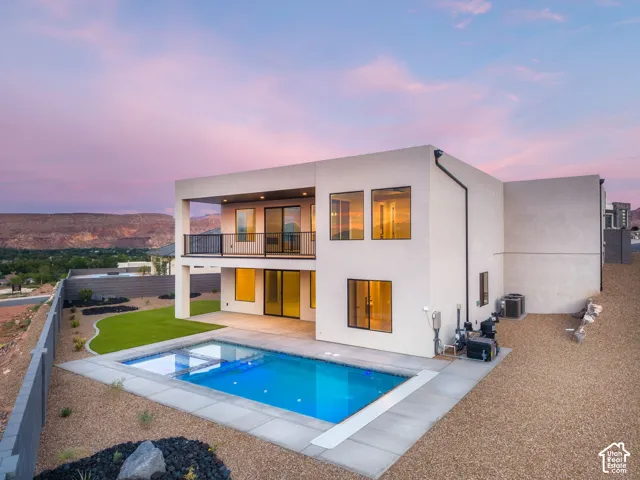 Modern architecture with a flat roofline, featuring an in-ground swimming pool, an upper-level balcony with metal railings, a lower-level patio, and a manicured lawn area