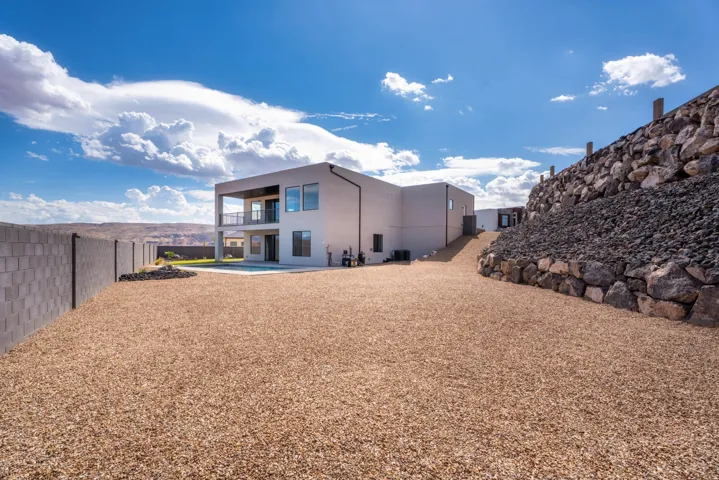 Modern stucco exterior with an upper-level balcony, a lower-level patio, a block privacy fence, and a gravel-covered yard