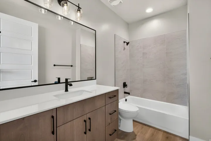 Wood-finish flooring, white solid surface countertop, wood grain vanity, black framed mirror, and tiled bath surround