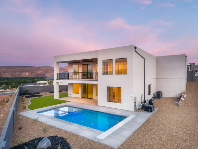 Modern architecture with a flat roofline, featuring an in-ground swimming pool, an upper-level balcony with metal railings, a lower-level patio, and a manicured lawn area