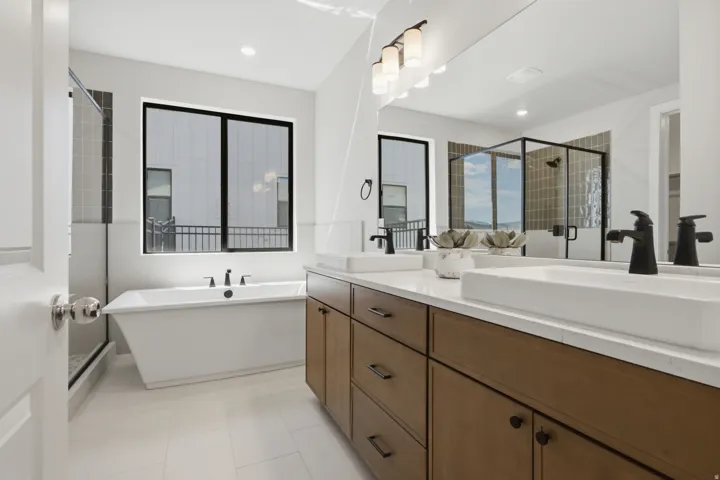Freestanding soaking tub with black fixtures, dual vanity with wood-finish cabinetry and rectangular vessel sinks, walk-in shower with clear glass enclosure and tiled walls, and large window with black trim